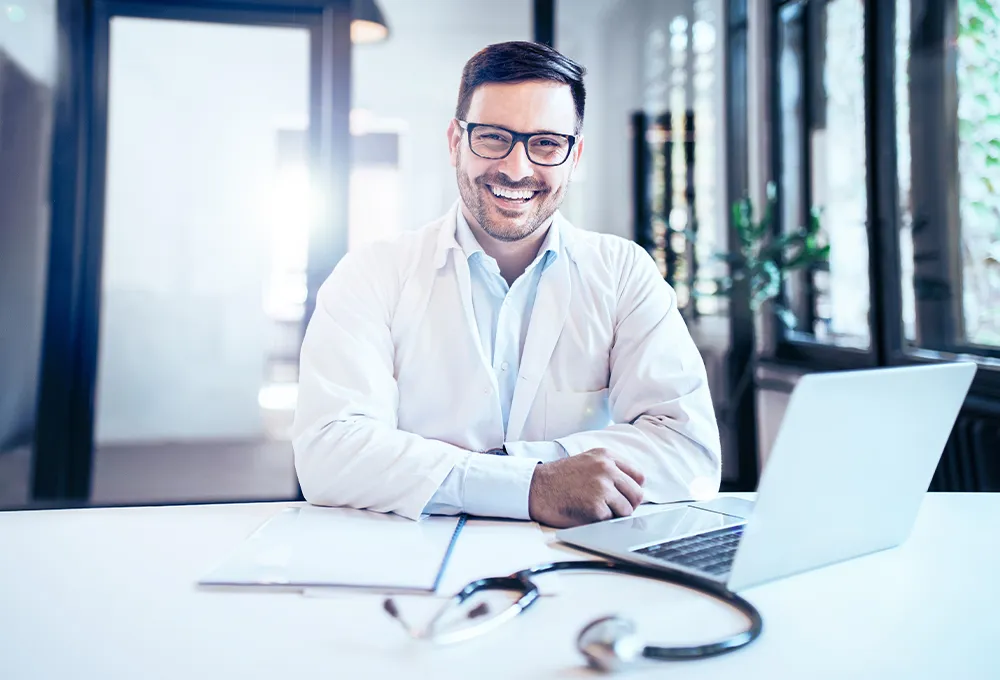 Arzt lachend und freundlich, sitzt am Tisch mit Laptop und Stethoskop